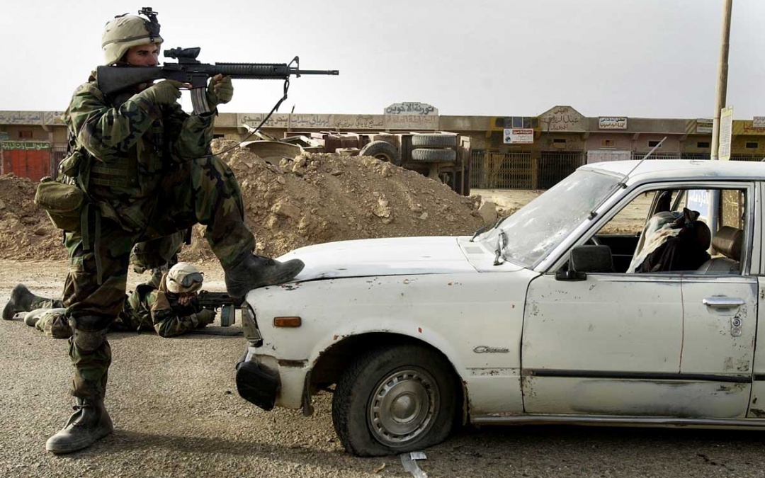 A Marine holds a rifle, positioning himself on the hood of a white care