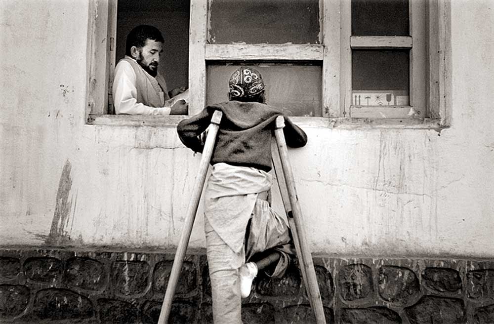Afghan boy with crutches who was a landmine victim stands at a window listening to a man read to him.