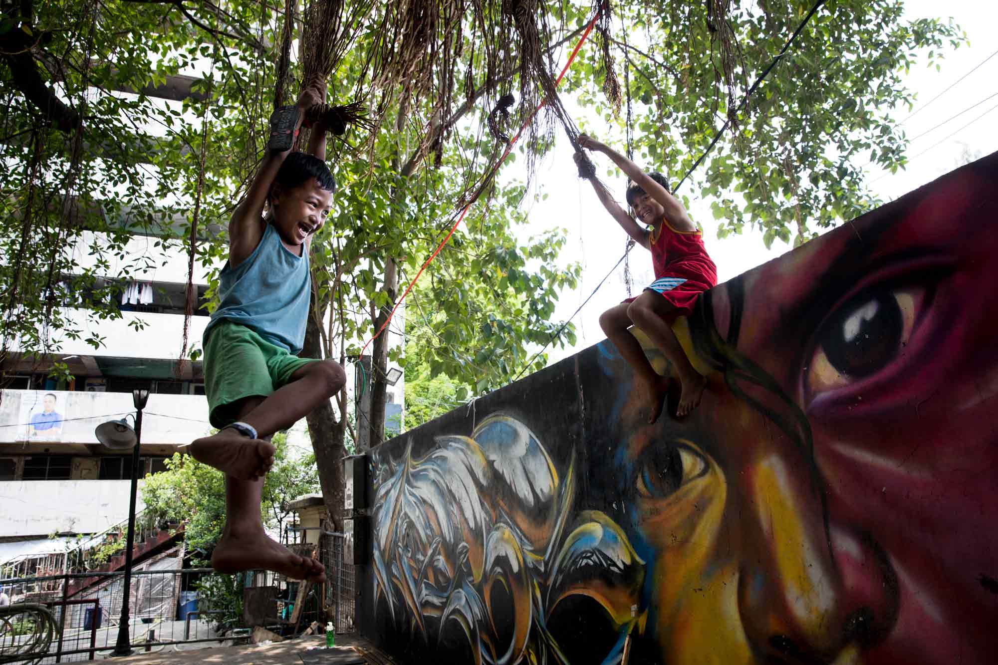Prince Jay Dadulla, 8, and Jaylord Diorico, 10, left and right, swing from vines that hang from an old tree in the public square at the Tenement in Taguig, Philippines. Many families residing at the Tenement have lived there for generations, entrenched in poverty.
