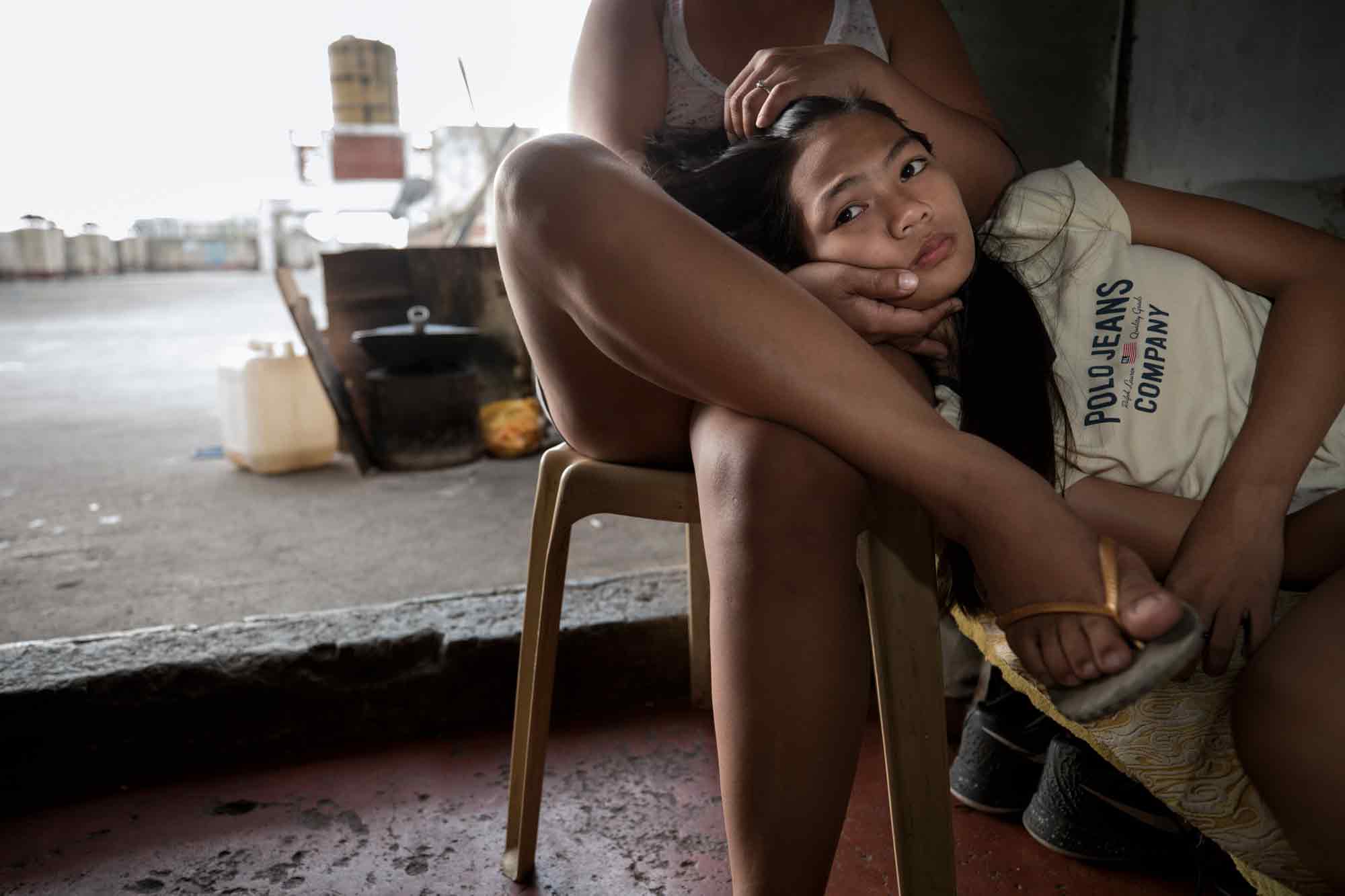 Michelle Nicdao, 14, lays in her mother Maria Oliva Pagayanan's lap in their rooftop unit which they share with her three brothers at the Tenement in Taguig, Philippines. In her youth, Pagayanan entertained men in nightclubs; it was her father who got her the job.