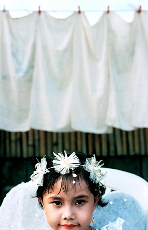 She waits by the dusty road, anxious to play her part as an angel in the Santacruzan procession so she can give honor to her family. Katrina Mayor's mother paid for a professional beautician to paint her five-year-old daughter's face and nails so that she might be more perfect in the eyes of God. Philippines A little girl wearing a headdress is framed by laundry as she readies for a Santacruzan procession in the Philippines.